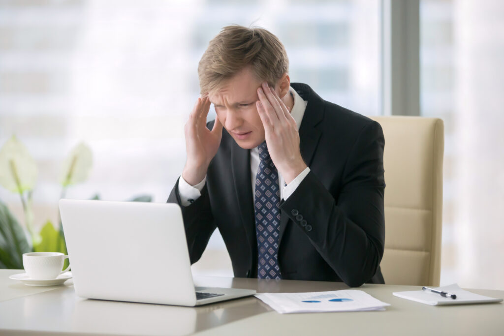 Young shocked businessman sitting with laptop at desk, having headache, poor cash flow, balance, low profit, unable to deal with fears, burning out, sacrificing physical and mental health for business