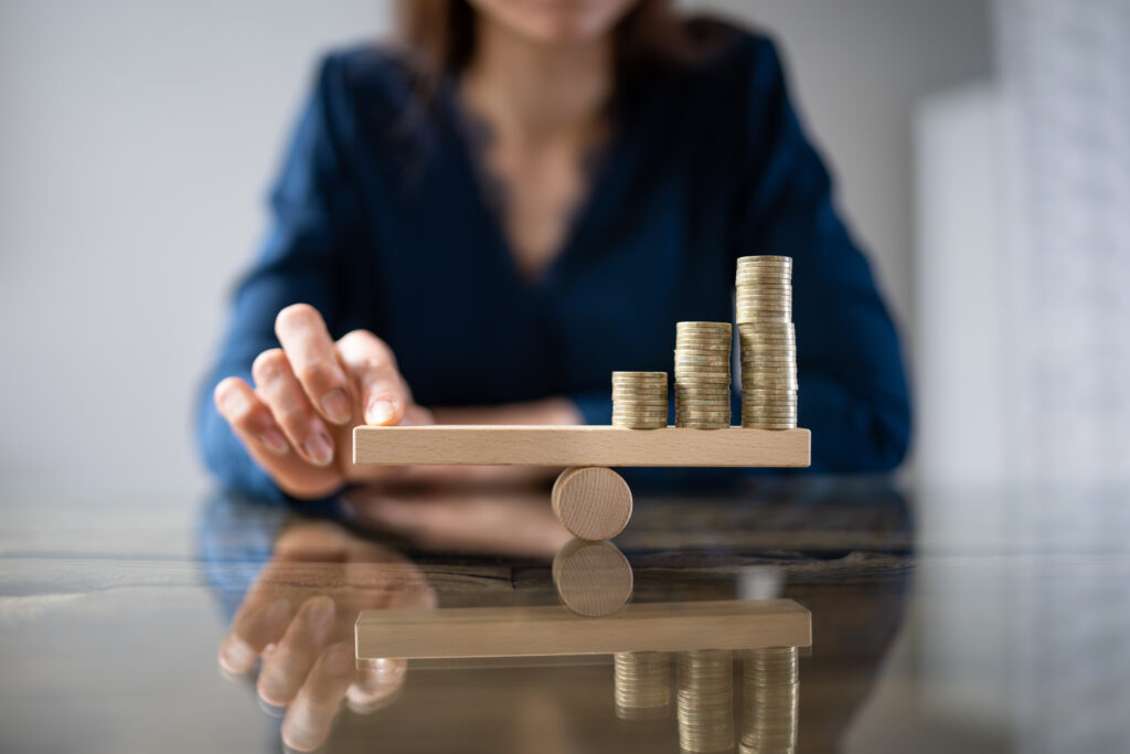 Close-up Of A Businessperson Balancing Stacked Coins On Seesaw With Finger